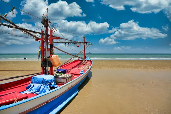 A red and blue traditional fishing boat on the sandy beach at Hua Hin beneath a cloud-filled sky