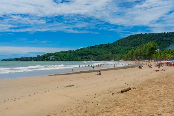 People swimming and relaxing along a sandy Phuket beach backed by palms and a forested hillside