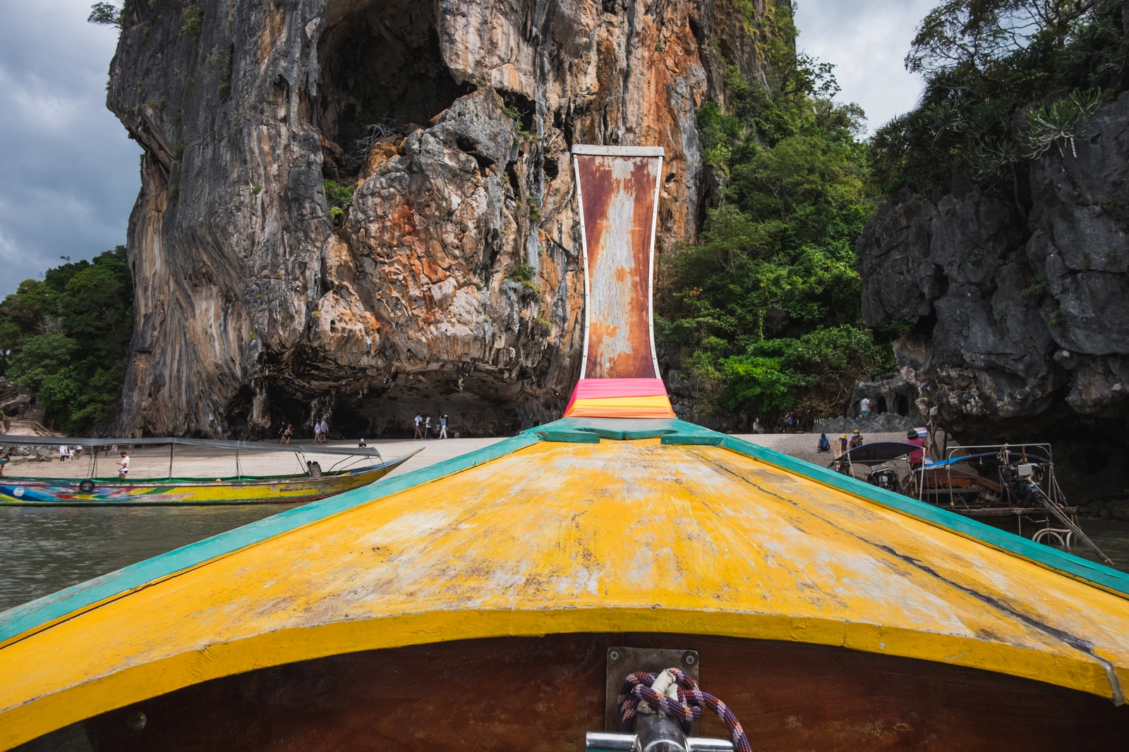 View over the yellow deck and bow of a long-tail boat toward a limestone cliff and beach